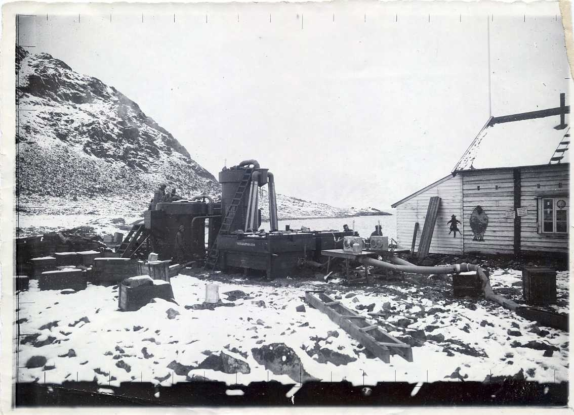 Vätgasapparaten Danskön, män i regnplagg står intill. Pikes hus till höger. Snö på marken.