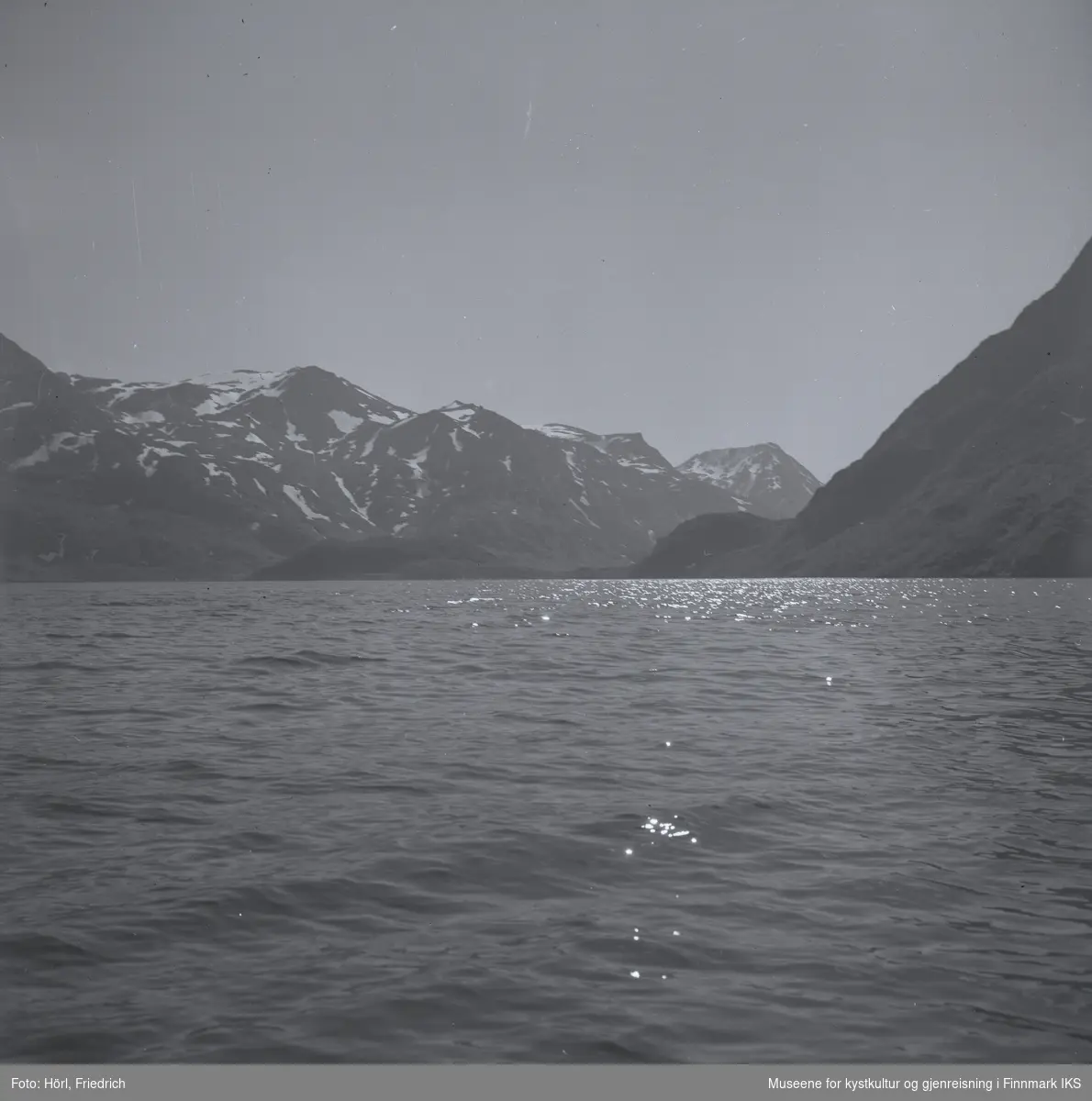 Motivet er fotografert på en båttur fra Hammerfest til Seiland. Sola glitrer på havets bølger og på fjellene liger det fortsatt noe snø.