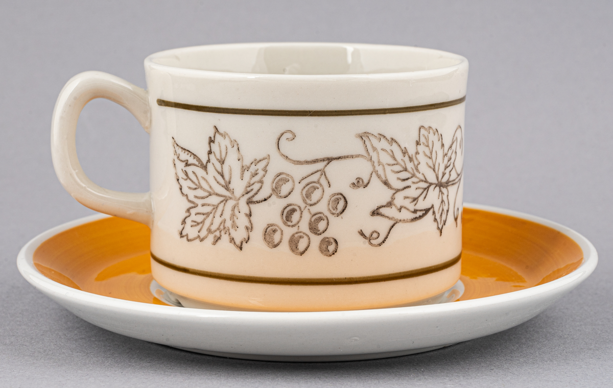 Kaffegods i flintgods, modell EH tillverkad 1971 och framåt. Okänt vem som skapat modell och dekor. Koppartryckt bladranka med handmålad rand upptill och nedtill på koppen, fatet med handmålad gulfärgat bräm.
