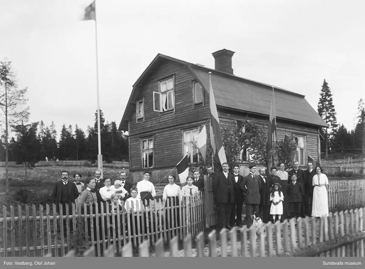 Gruppbild med vuxna och barn framför ett bostadshus med brutet tak, en stor yta är inramad med spjälstaket. Flaggan är hissad och grindpassagen är lövad och omgiven av olika flaggor.