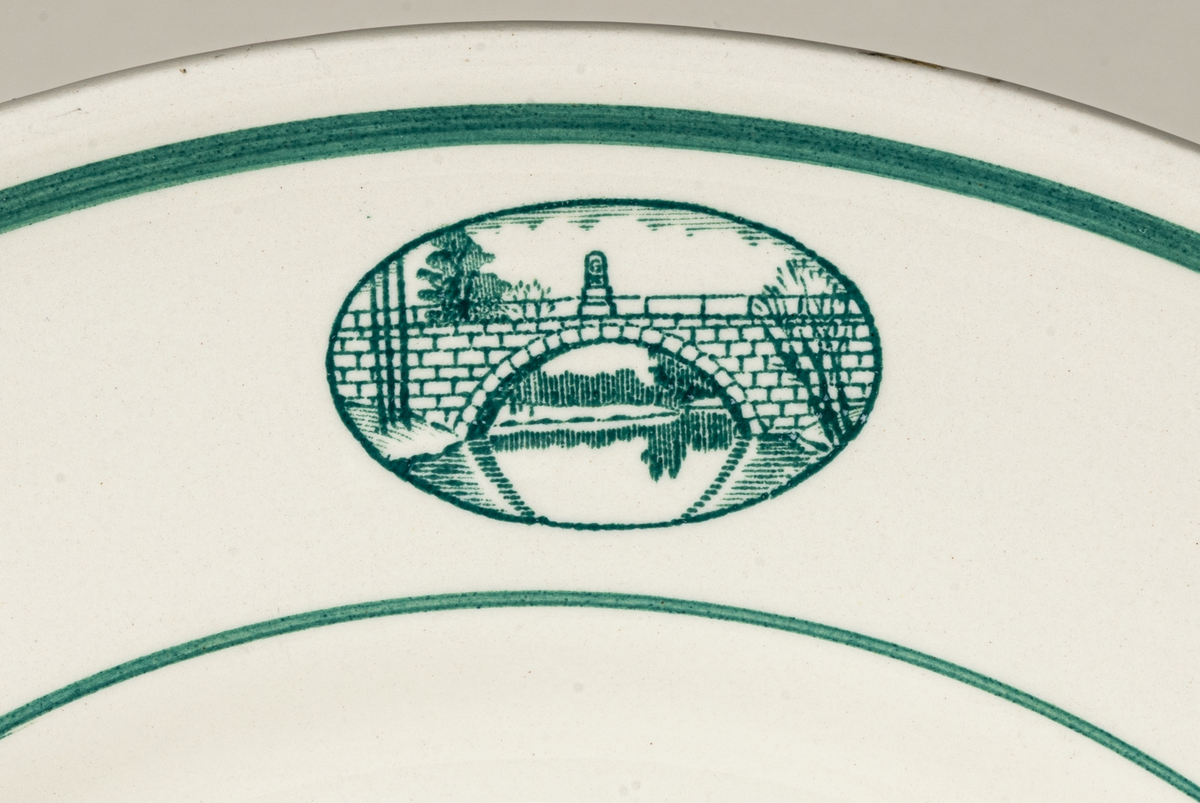Flat tallrik till matservis med bild/logotype på brämen föreställande Gustavsbro vid Gävles västra utfart, en bro över Gavleån som byggdes 1782. För vem och varför servisen tillverkades är okänt. Dekoren av två linjer runt brämen samt logotypen är i grönt.