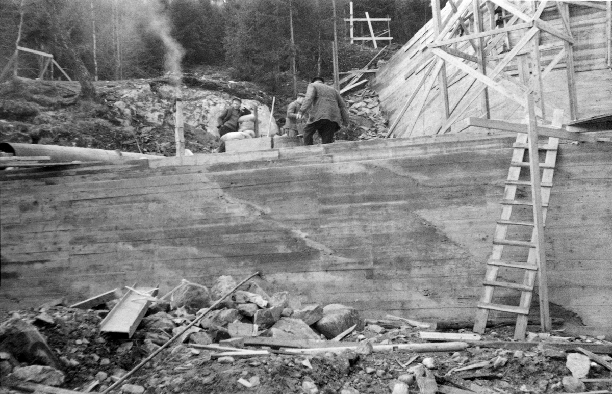 Fra bygginga av en ny dam ved Sølvstufossen, øverst i Ågårdselva i Tune i Østfold, som er en del av Nedre Glommas vestre løp. Anleggsarbeidet startet vinteren 1936, i en periode da det knapt rant vann i elveløpet. Dette fotografiet er tatt noe seinere, etter at snøen i terrenget hadde smeltet bort, og etter at mye av murverket var ferdigstilt og etter at bordforskalingene var revet. Over damkrona sentralt i bildet ser vi likevel tre arbeidere i aktivitet ved det dampdrevne betongblanderiet. Ågårdselva er et cirka fem og en halv kilometer langt vassdrag som renner fra den nordvestre enden av Isnesfjorden (Vestvannet) i Nedre Glommas vestre løp, sørvestover gjennem berglendt terreng mot innsjøen Visterflo. Høydeforskjellen mellom Isnesfjorden og Visterflo er på bortimot 25 meter. Ågårdselva har tre fossefall, det øverste her ved Sølvstu, deretter ved Valbrekke og nederst ved Solli. Behovet for den dambygginga vi ser på dette fotografiet var forårsaket av selskapene Borregaards og Hafslunds inngrep i Glommas østre løp, hovedløpet, som i lavvannsperioder gjorde det svært vanskelig å få tømmer som skulle til bedrifter nedenfor Sarpsfossen via Mingevannet, Isnesfjorden og tømmertunnelen til Eidet gjennom det trange sundet ved Trøsken. Dette forsøkte man å løse ved å heve vannspeilet i Isnesfjorden med en dam som skulle plasseres 10-15 meter ovenfor den eksisterende dammen ved Sølvstufossen. Se mer informasjon under fanen «Opplysninger».