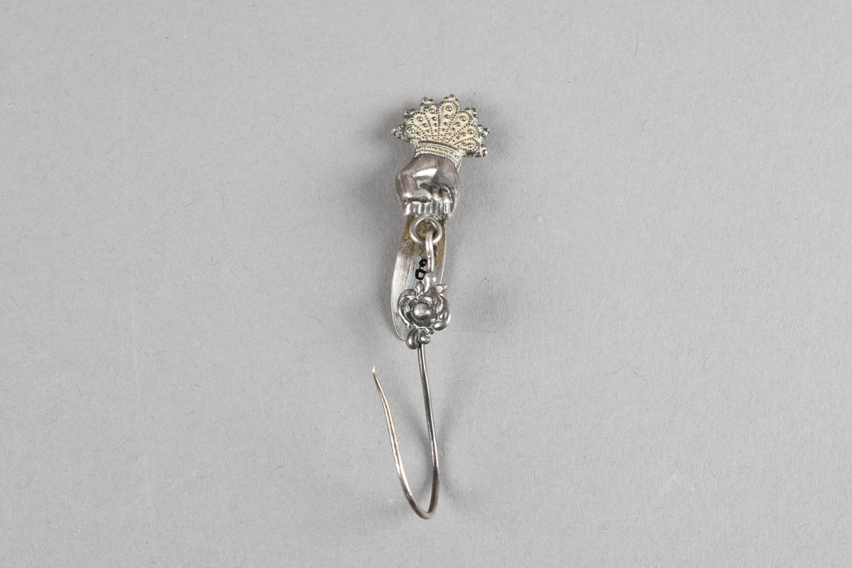 Nøstekrok av sølv. En knyttet hånd med kroneformet mansjett over. Hånden holder en bøyle som er formet til en krok.