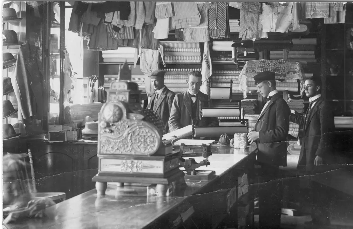 Interiör från konsumbutiken på Bruksgatan som öppnade 15 april 1908. Butiken var belägen mellan Bruksgatan 5 och 7 och ägdes av pappersbruket. Verksamheten flyttade från Bruksgatan till det nybyggda huset vid Nyboda 1911. Husmödrar i kvinnogillet sydde upp skjortor som såldes i butiken.