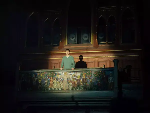 En mann står opplyst oppe på galleriet i en kirke, det er maleri av Jesus på galleriveggen foran mannen.