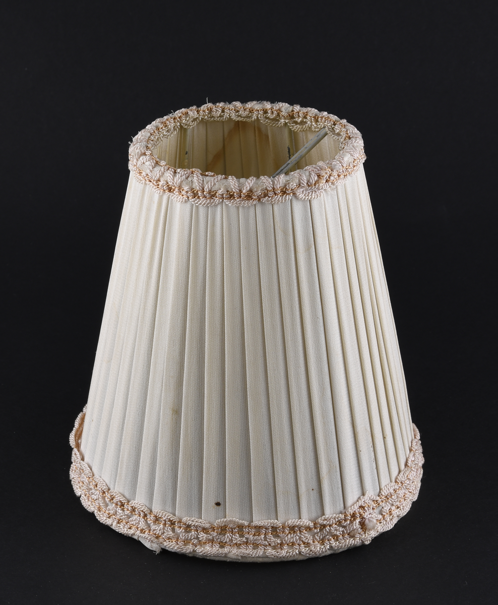 Lampeskjerm av lys tekstil. Konisk form, med dekor i topp og bunn av flettede tråder.
