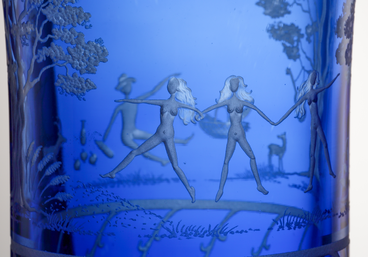 Formgivare: Gunnar Cyrén. Vas med bägarform, blå fond. På livet graverad dekor föreställande ett naturmotiv med 
olika människofigurer och djur. Graverad bård på vasens nedre del.