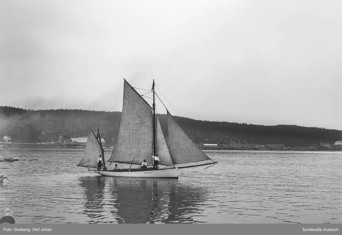En segelbåt med fyra unga män ombord, mellan Essvik och Svartvik. Bakom syns Svartviks sågverk med herrgården längst till vänster och längst till höger skymtar missionshuset i Vapelnäs.