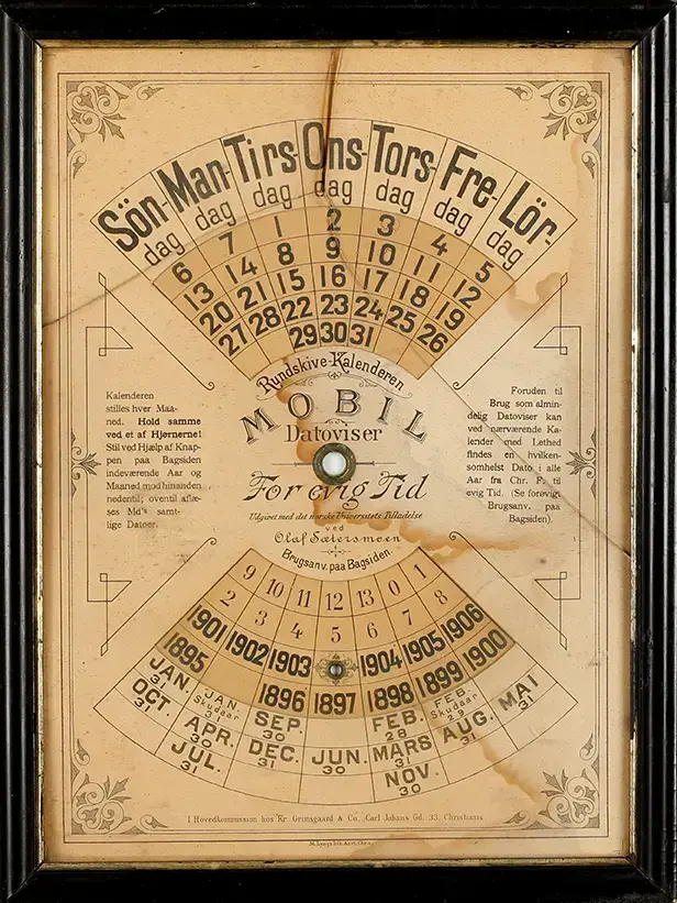 En avlang evighetskalender i papp i en mørk treramme. Kalenderen har en rund skive som kan dreies for å vise korrekt dato. Fint dekorert med grafiske elementer fra tidlig 1900-tall.