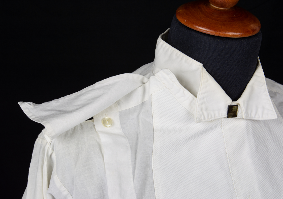 Vit stärkskjorta med snibbkrage, stärkt framstycke samt
dubbla manschetter för manschettknapp. Skjortans knäppning sitter på sidan om framstycket upp mot axeln. I kragens knapphål sitter en löstagbar fyrkantig knapp av plast.