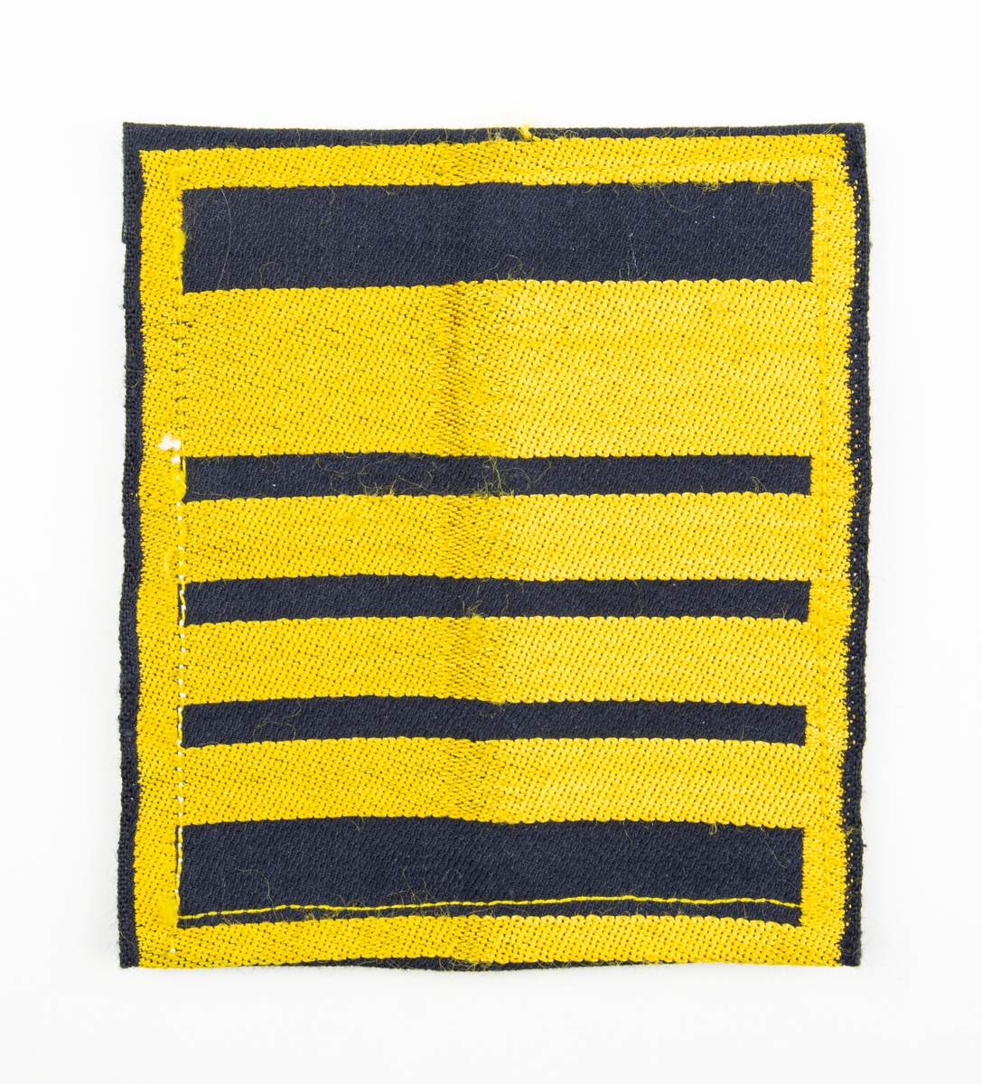 Ärmmatta, överste, med invävd gradbeteckning i gult på svart botten. På baksidan fastsydda kardborreband.