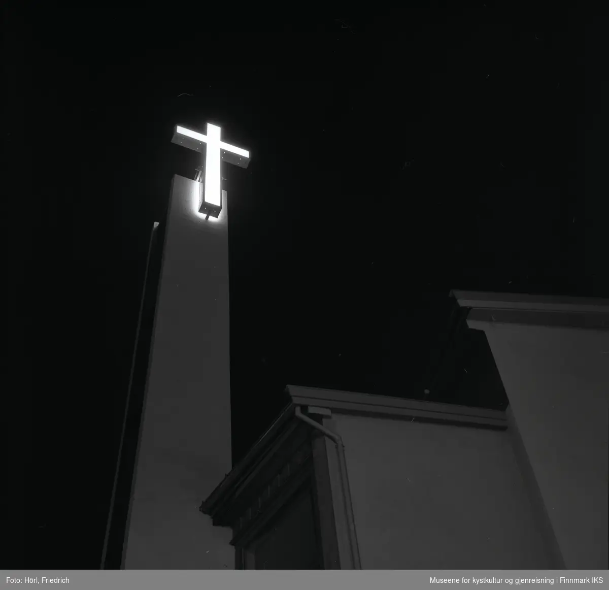 Det lyssatte korset ved klokketårnet til den katolske kirka St. Mikael i Hammerfest lyser i nattemørke. Bildet er tatt oppover fra gateplan i Strandgata.