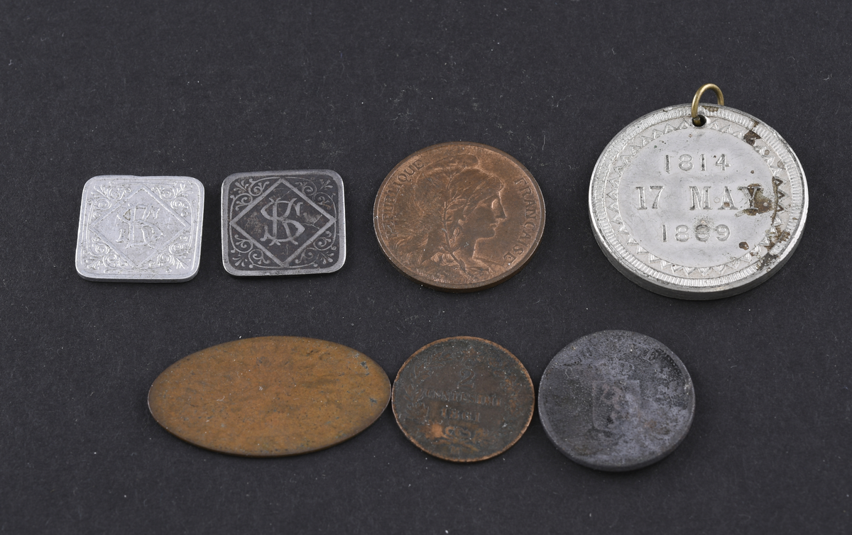 Syv mynter, medaljer og polletter i varierende størrelse og tilstand. Myntene er fra USA, Tyskland og England. Myntene og polettene var oppbevart i en liten papirpose.