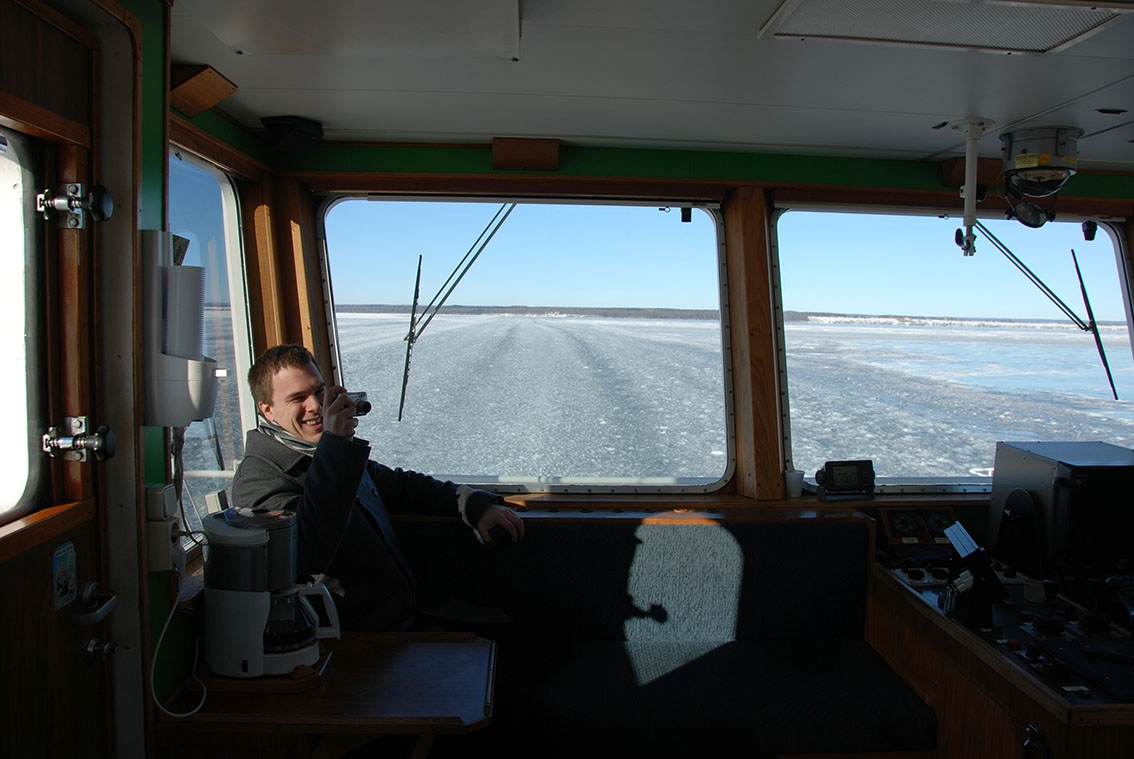 En man sitter i kommandobryggan på färjan från Gränna till Visingsö. Han fotograferar troligen befälhavaren. Genom fönstret ser man rännan i isen.