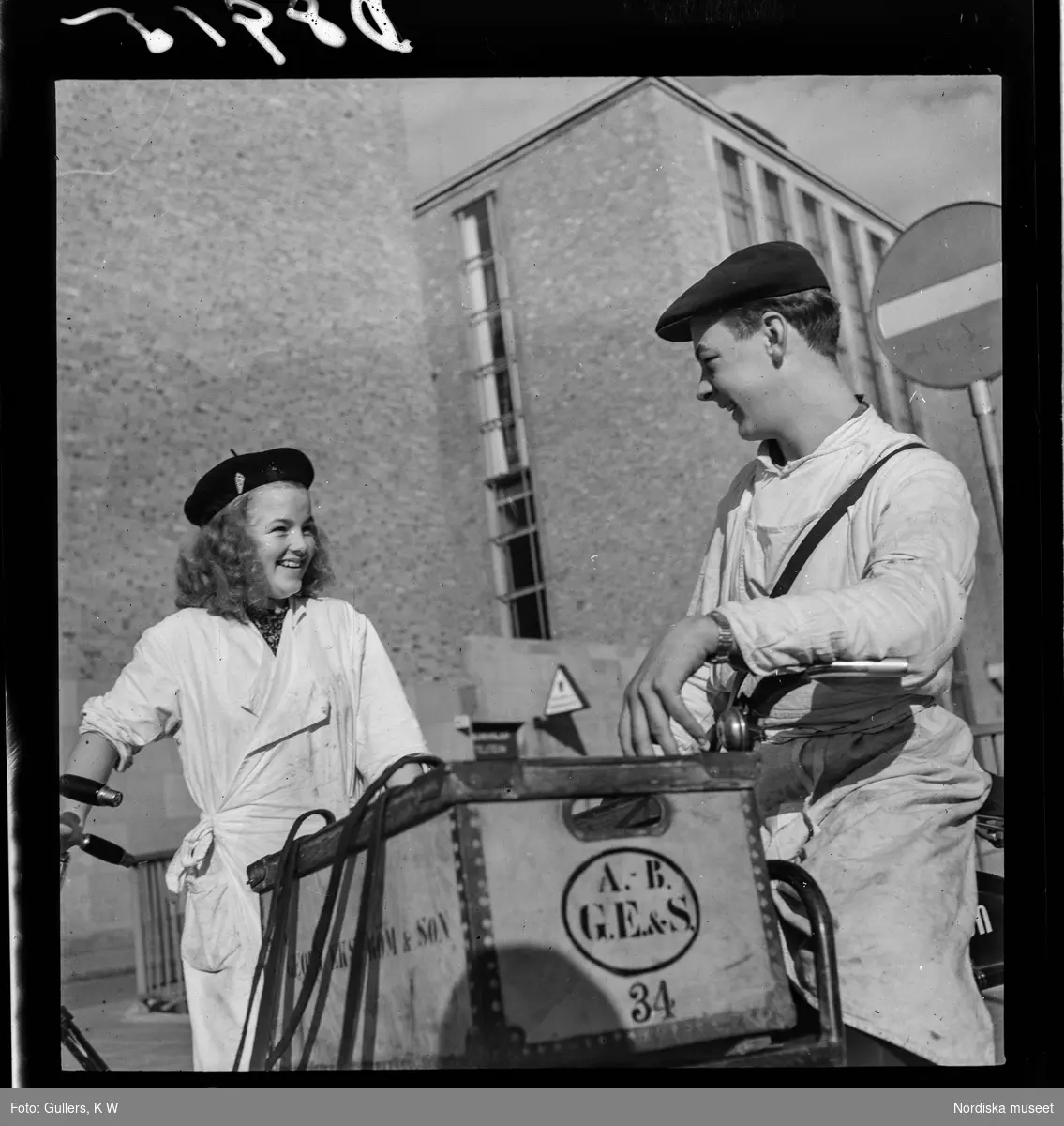 539 Söder. En ung kvinna och en ung man i arbetskläder på cyklar. Mannen har en låda fastspänd framtill på cykeln med texten Georg Ekström & Son.
