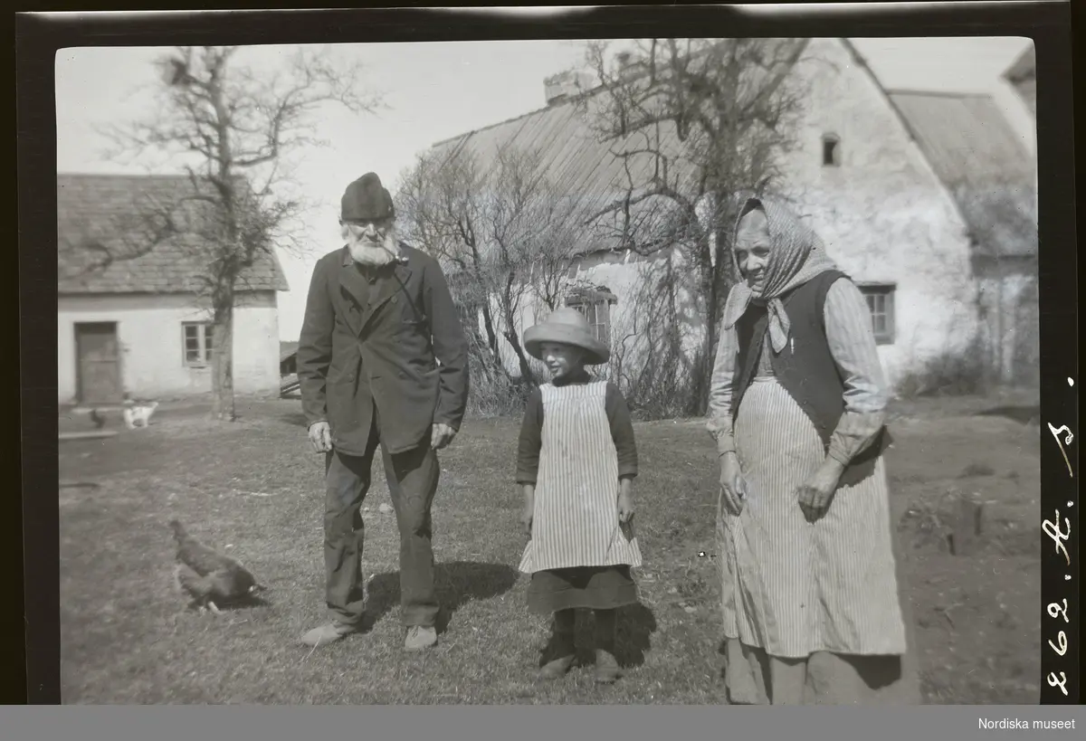 En äldre man, en flicka och en äldre kvinna utomhus på en gårdsplan.