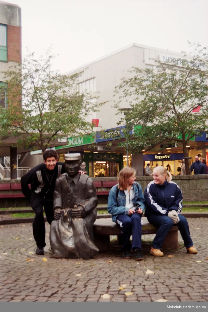 Tre ungdomar vid skulpturen Albert på Mölndals torg. Mölndalsbro i dag - ett skolpedagogiskt dokumentationsprojekt på Mölndals museum under oktober 1996. 1996_1190-1211 är gjorda av högstadieelever från Kvarnbyskolan 9D, grupp 4. Se även 1996_0913-0940.