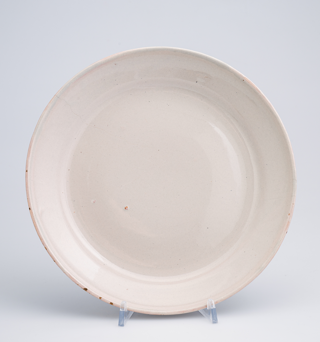 En flat tallerken av keramikk/porselen. Den har bly- og tinnglasur og er hvit på farge. Tallerkenen er rund med en relativt høy og skrånende kant. Den har ikke dekor utover glasuren. Tallerkenen er en av 11 identiske tallerkener som trolig er fra tidlig på 1700-tallet.
