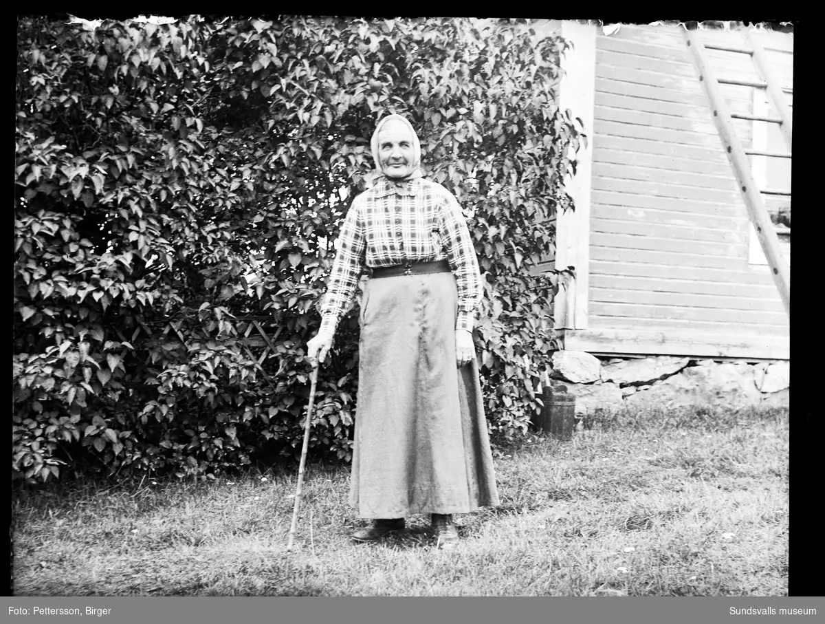 Porträttbild av en äldre kvinna med käpp ute i trädgården, en husfasad syns i bakgrunden.