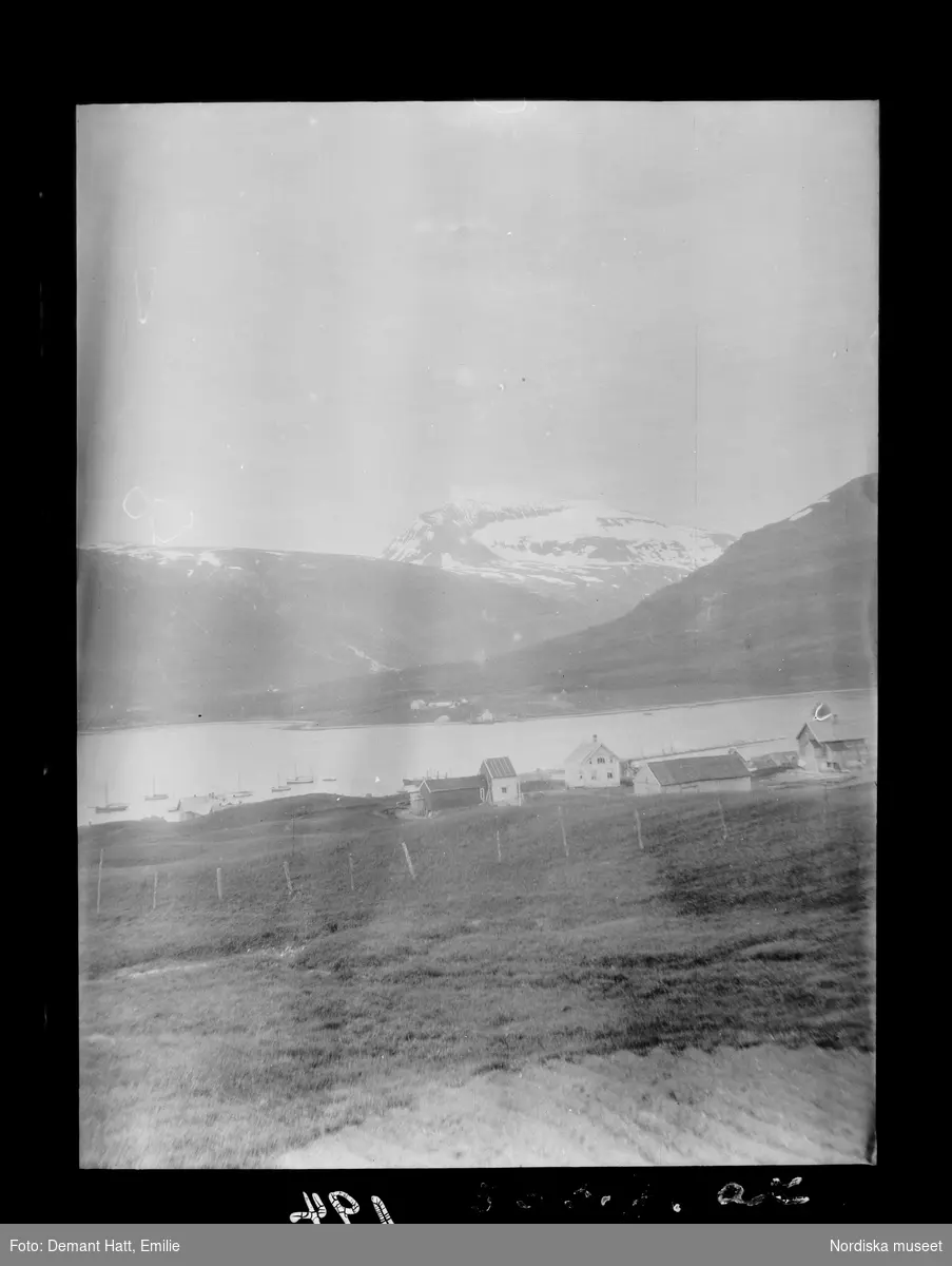 Vy över bebyggelse, åkrar, vatten och fjäll i Norge. Bygden som idag är en förort till Tromsø heter Hungeren. Bilden ingår i en serie fotografier tagna av Emilie Demant Hatt i Sapmi mellan åren 1907 och 1916.