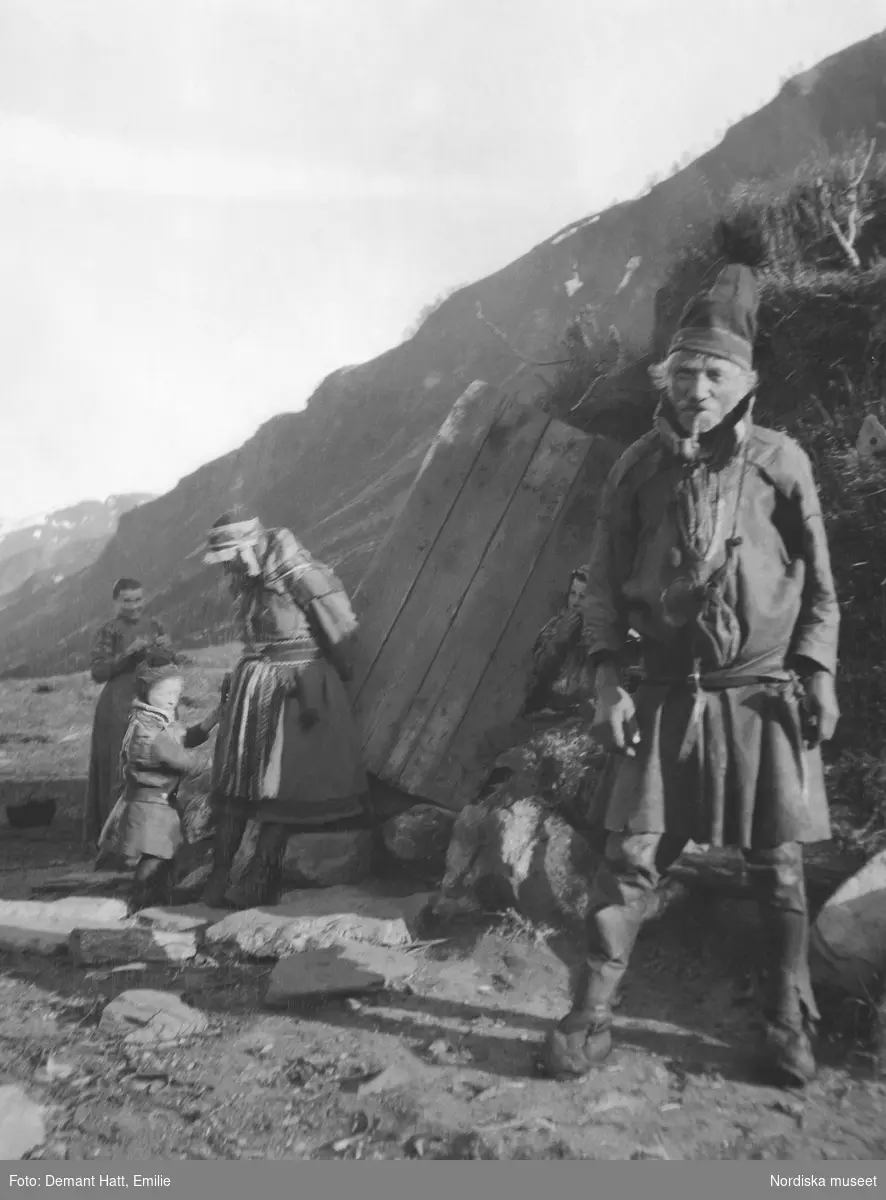Framför en kåta står en man, Hoalsta-Lasse. bakom ses kvinnor och barn. I bakgrunden fjäll.  Bilden ingår i en serie fotografier tagna av Emilie Demant Hatt i Sapmi mellan åren 1907 och 1916.