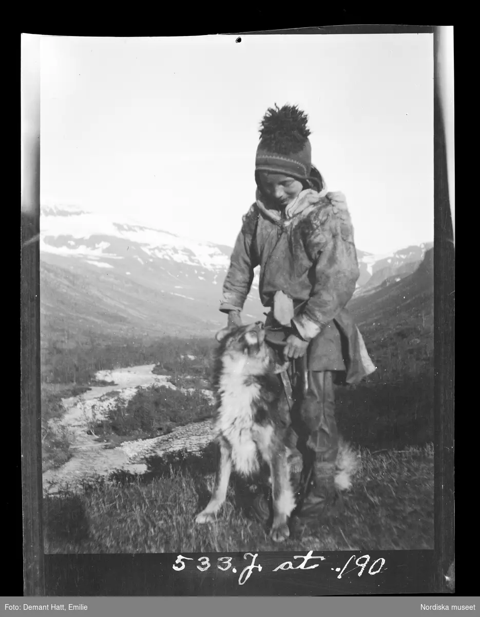 Ett barn leker med en hund (Curri), enligt bildtexten är pojken Andaras Ommas yngste son. I bakgrunden fjäll och en dalgång. Bilden ingår i en serie fotografier tagna av Emilie Demant Hatt i Sapmi mellan åren 1907 och 1916.