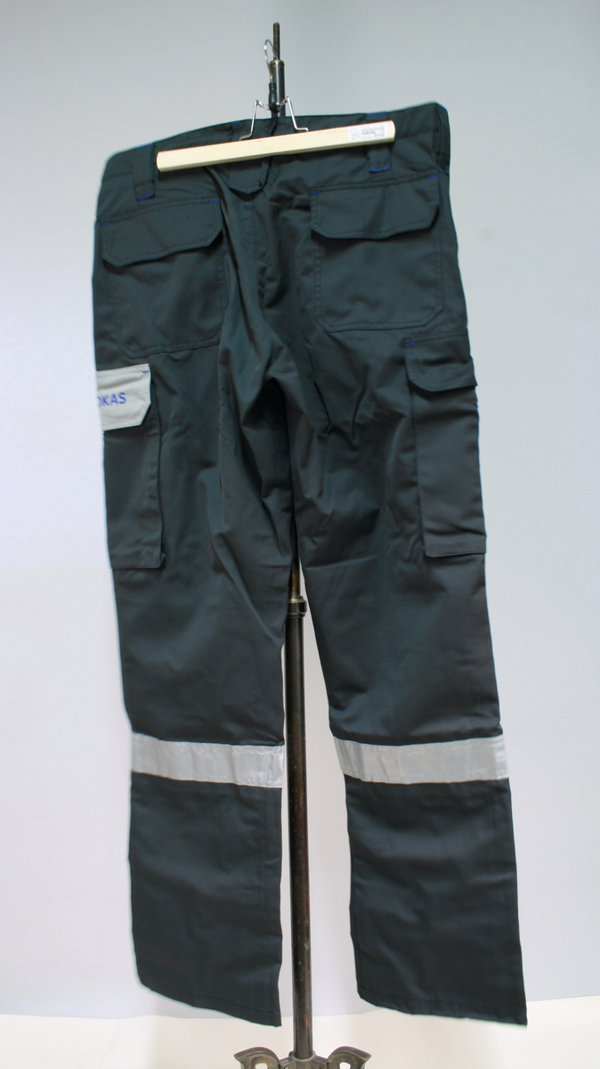 Sort uniform bestående av kortermet poloskjorte, bukser og skyggelue med Nokas-logo, og tilhørende belte rigget med lommelykt og walkie-talkie.