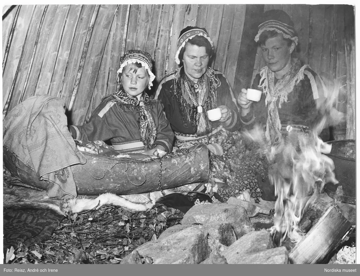 Samer, Norrland. Interiör av en lappkåta. Två kvinnor och en flicka, i samedräkter, sittandes med en samisk vagga vid den öppna elden.