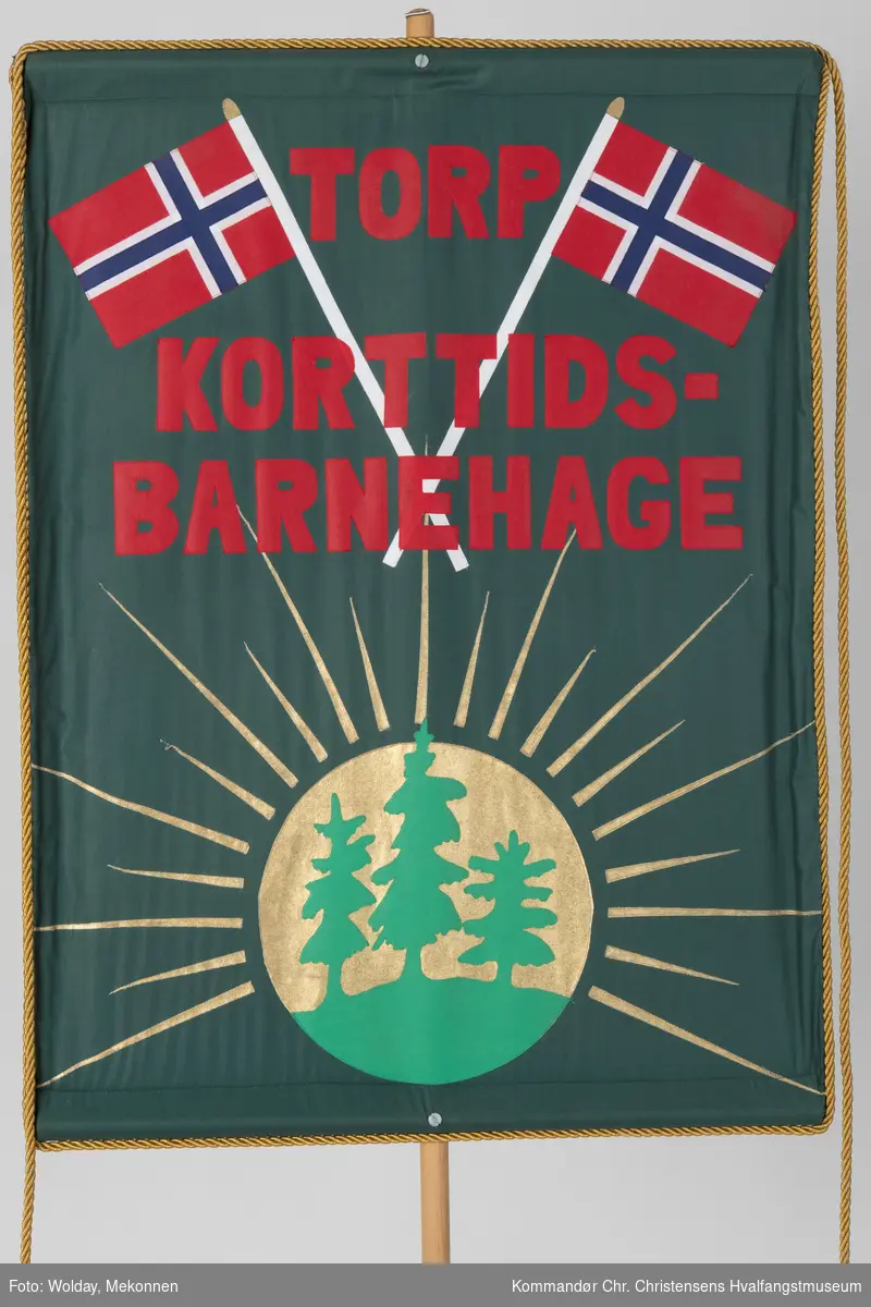 Mørkegrønn bakgrunn med rød skrift. Dekortert med norske flagg og en sol med grantrær. Festet til en rundstokk.