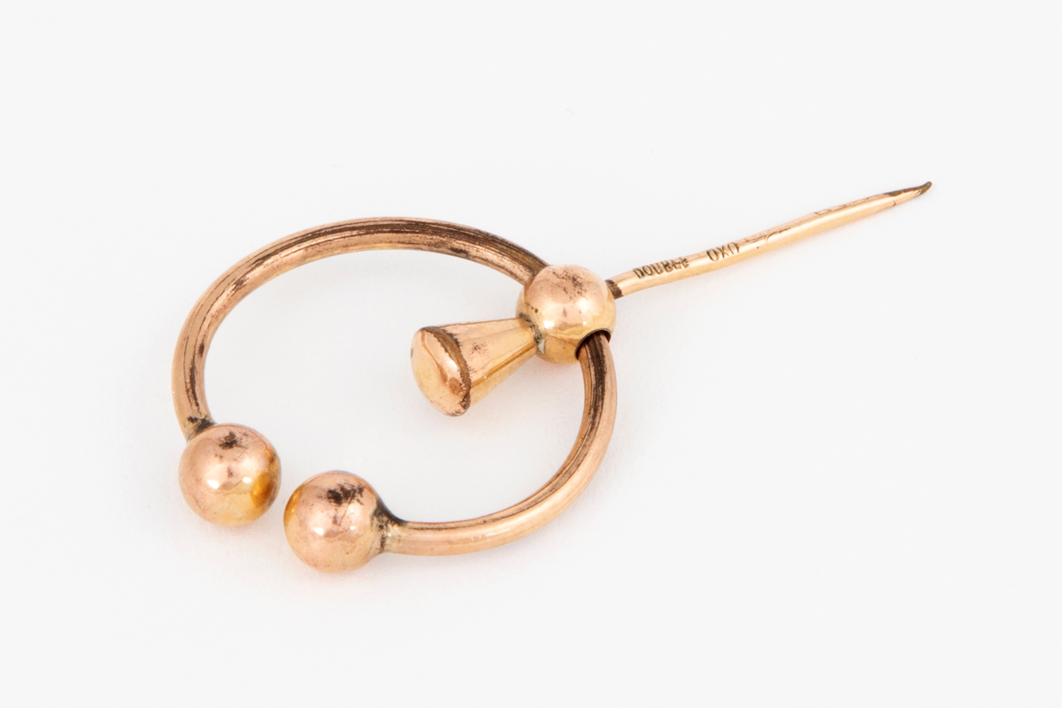 Brosje i gulldublé formet som en åpen ring med to kuler i hver ende. En bevegelig nål med kjegleformet hode er festet til ringen med en tilsvarende kule.