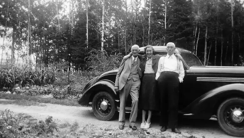 To eldre menn står med en kvinne mellom seg foran en mørk bil. Kvinnen har armene rundt mennenes skuldre. Bak bilen til venstre er en grønnsakshage med noen trær bak.