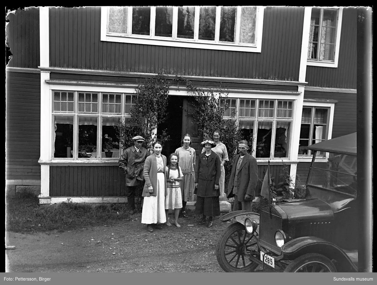 Gruppfoto vid den lövade ingången till en glasveranda, kvinnor, män och en flicka samt även en bil med registreringsnummer Y285. Bilen är en T-Ford från cirka 1923-25.