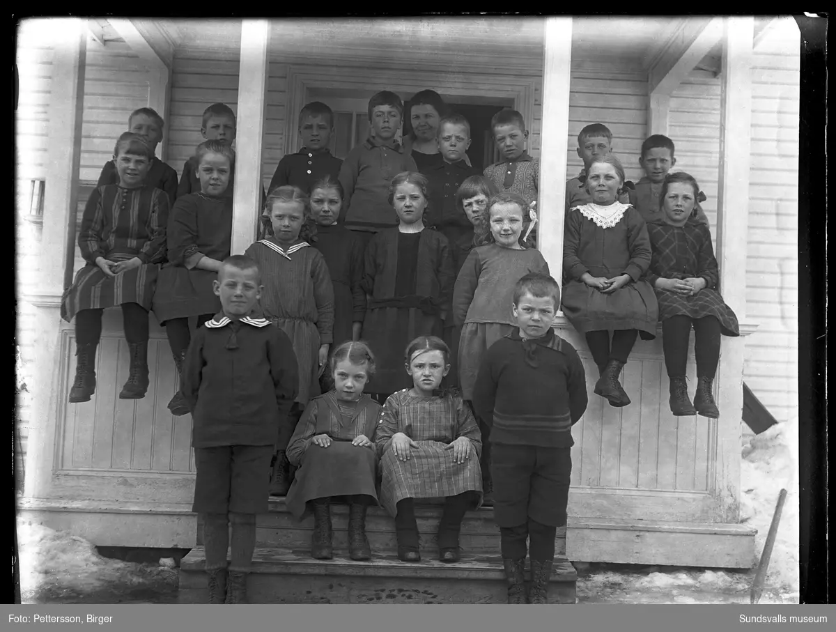 Gruppfoto med barn på brokvisten till ett ljusmålat hus, sannolikt ett skolfoto. Längst bak en kvinna/lärarinna. Samma brokvist och kvinna som SuM-foto035239.