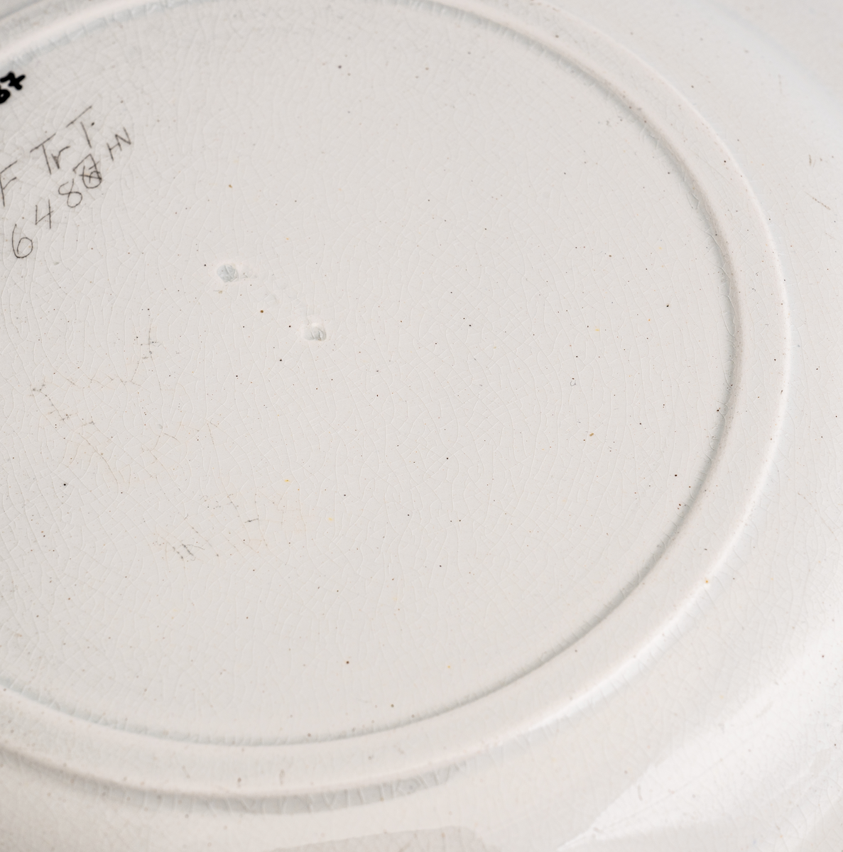 En mellomstor flat tallerken av glasert keramikk/porselen. Den er hvit på farge (ensfarget). Kanten er inndelt i 12 felt "adskilt" med loddrette tversgående streker som stikker opp/som er støpt i kanten. Flaten på midten har en diameter på 16,5cm. Det er inskripsjon/dypstempel på undersiden.
