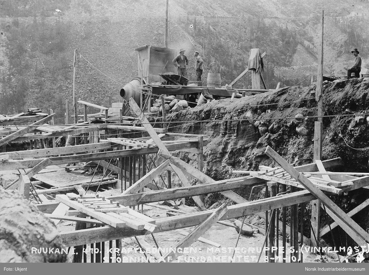 Støping av fundament for mast til kraftledningen på nordsiden av dalen mellom Vemork og Rjukan. Arbeidere på anleggsområdet. Vemorksporet i fjellsiden i bakgrunnen.