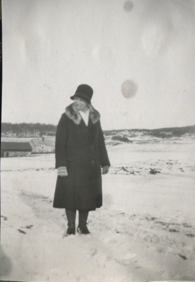Svea Johansson står vinterklädd med kappa och hatt i ett snörikt landskap, Heljered Mellangård på Hållevägen, okänt årtal.