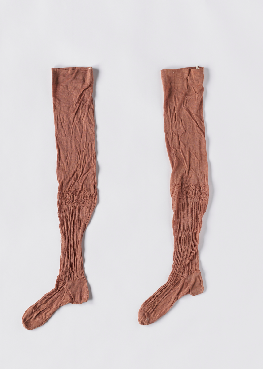 Strikkede rosa strømper i bomull. Foten er formsydd. Det er strikket hullmønster fra oversiden av foten og oppover leggen. Det er hull i brettekanten for å tre inn bånd.