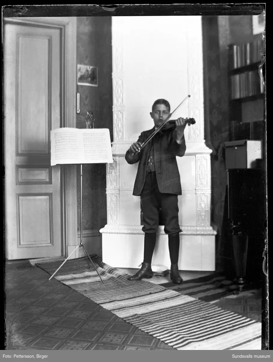 En pojke står framför en kakelugn och spelar fiol. Bredvid står ett notställ.