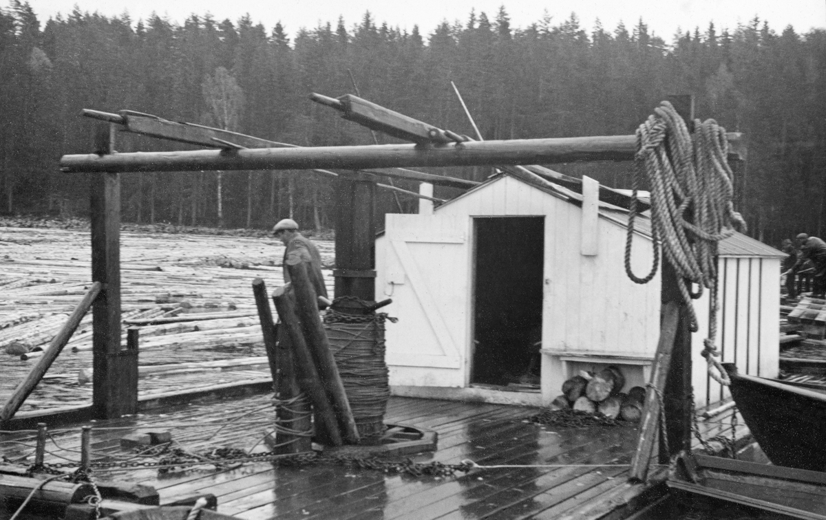 Spillflåte på Hurdalssjøen i Akershus, 1939.  Fotografiet er tatt på nært hold, mot det bordkledde dekket på flåten.  Sentralt på farkosten er det montert et gangspill med ei tønne med tauverk.  Gangspillet er hengslet i flåten og i en horisontal bjelke, som kviler på stendere ved flåtens ytterkanter.  Oppå den horisontale bjelken ligger også et par årer, som da dette fotografiet ble tatt kvilte på taket på et bakenforliggende, kvitmalt bordskur med saltak. Et par robåter lå fortøyd på høyre side av flåten, mens tømmeret lå tett på vannspeilet til venstre i bakgrunnen.