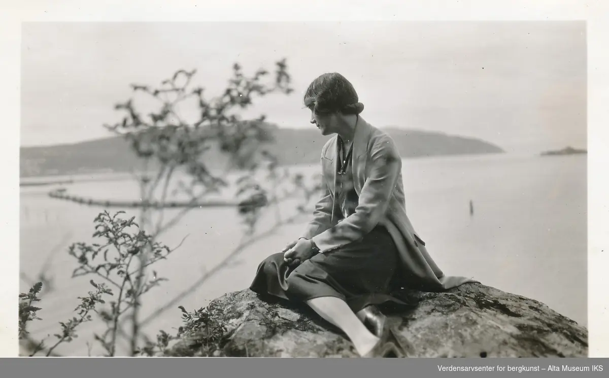 En kvinne sitter på en stein foran sjøen, hun ser vekk fra kameraet og har hendene holdet i fanget. Kvinnen er i fokus, og landskapet bak og tregrenen foran henne er ufokusert. 