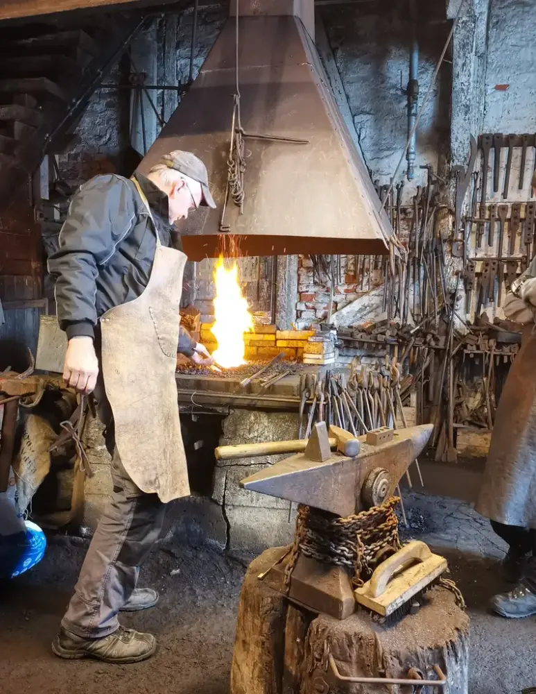 En eldre mann står i en historisk smie. Han har på seg et skinnforkle og har ryggen vendt mot kamera og rører i kullet i smia. Kullet brenner med høy flamme