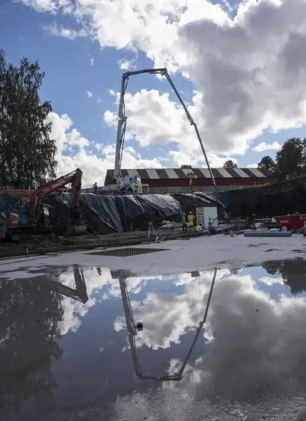 En betongbil med høy arm med slange står i skarp silhuett mot blå himmel med sol bak hvite skyer, speilbildet av det hele ligger i en vanndam i nedre halvdel av bildet.