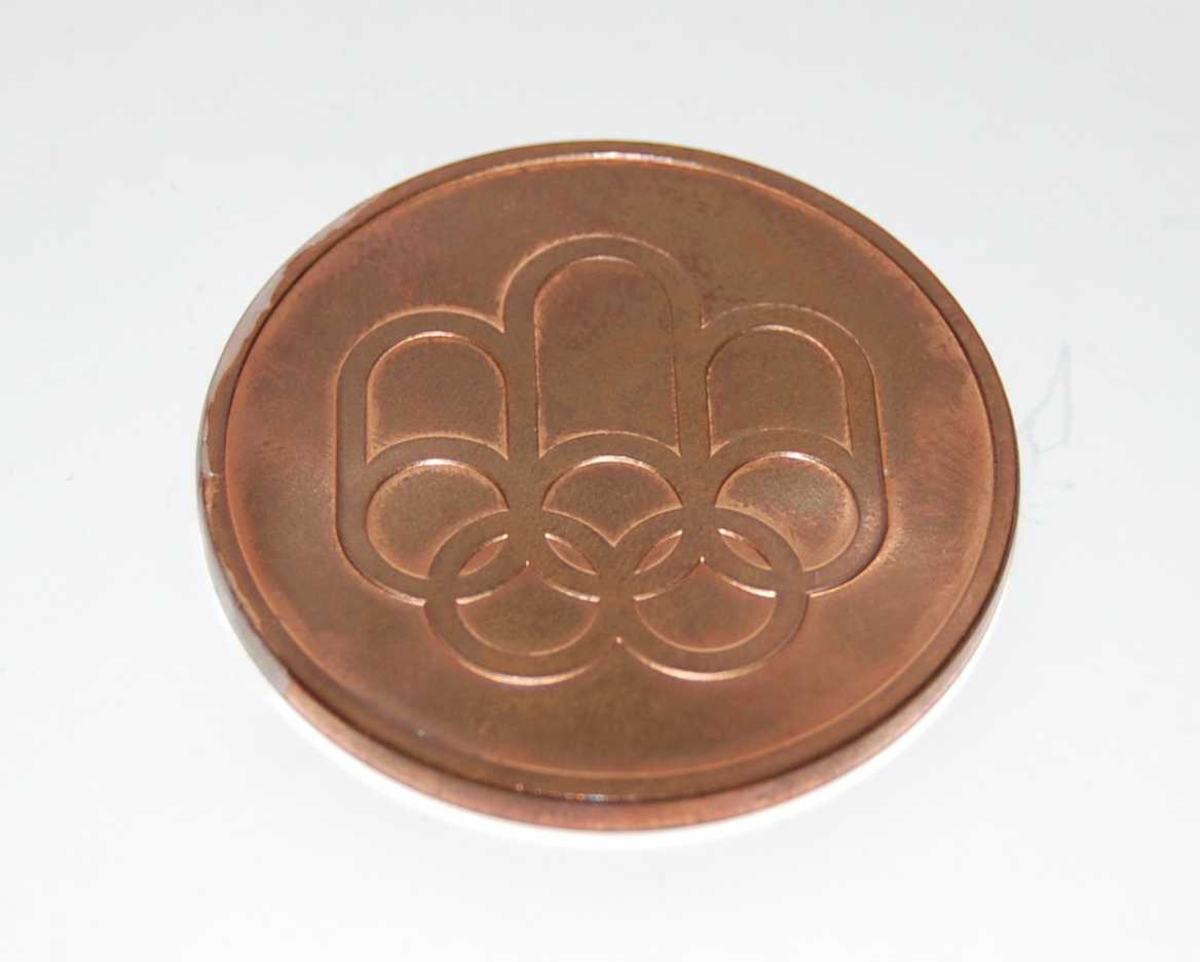 Bronsefarget minnemdalje med emblemet for de olympiske sommerleker i Montreal i 1976 og motiv av den olympiske stadion i Montreal. Med medaljen følger det et sort etui.