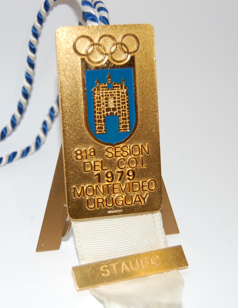 Gullfarget og blått merke med motiv av et våpenskjold preget av de olympiske ringene og en bygning. Til merket er det festet et hvitt bånd og en blå og hvit opphengssnor.