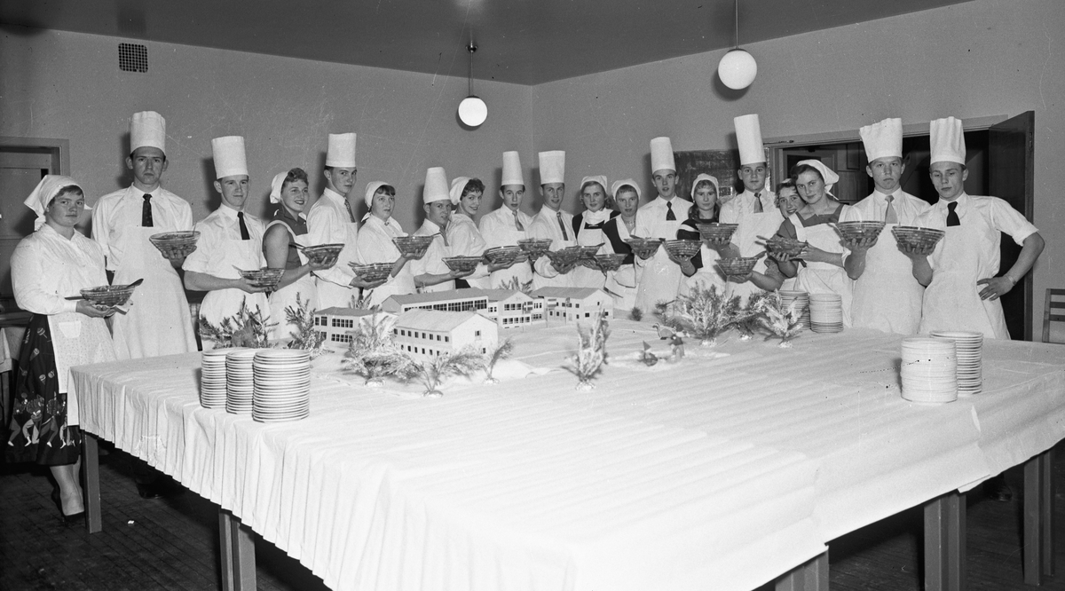 Stor gruppe elever i kokkeuniform. Modell av Romerike Folkehøyskole på bordet.