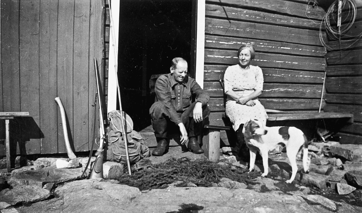 Kvinne og mann med jaktutstyr utenfor et tømmerhus.
