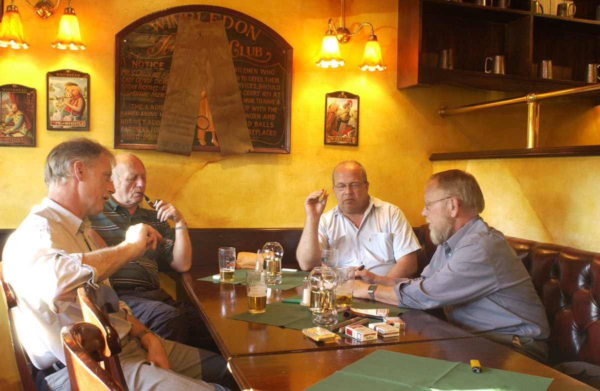 4 menn ved et bord med sigarer og pipe. Bowler & Spisepub.
