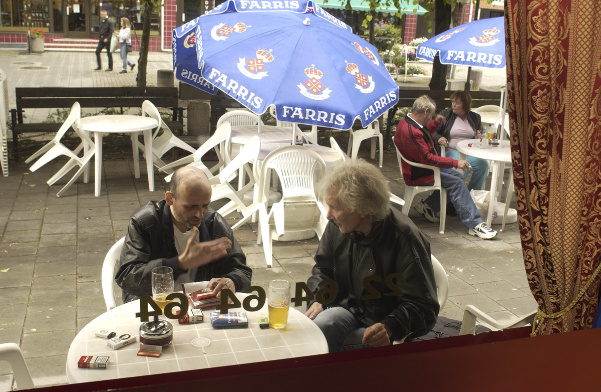 Første dagen etter innføring av røykeloven 2004. Flamenco Pizza & Pub. Parasoller og hagemøbler. To menn sitter ved et bord og røyker.