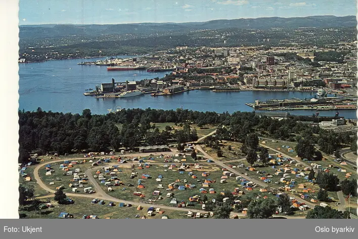 Ekeberg Camping ved Ekebergsletta, campingplass, telt, campingvogner, biler. Ekebergskogen. Panorama over indre Oslofjord og byen. Bispevika, Bjørvika, Vippetangen, Akershus festning, Kvadraturen. Postkort. (Flyfoto)