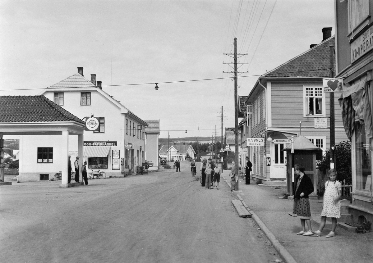 Postkort, Brumunddal med riksveg 50 gjennom sentrum. Standard bensin, Sandbakken bensinstasjon, O. Sønes, Brumunddal Bok & Papirhandel.
